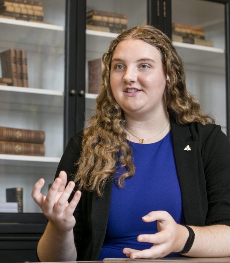 Annabelle looks cheerful while talking, in a black blazer over a blue dress, and long curly hair